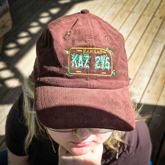 A person wearing a brown corduroy baseball cap with a 'KAZ ZY5' Kansas license plate patch on the front, photographed outdoors on a wooden deck in bright sunlight.